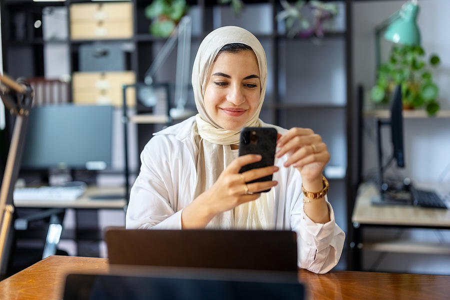 A young woman in a hijab smiles and uses her cell phone in an office environment