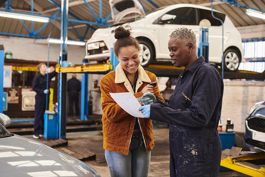 A woman and a mechanic in an automotive shop reviewing an estimate with a white car on a lift in the background.