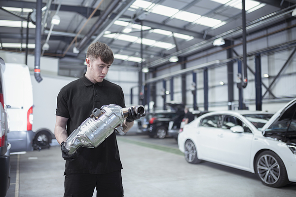 Man carrying a catalytic converter through an auto repair shop past a white sedan.