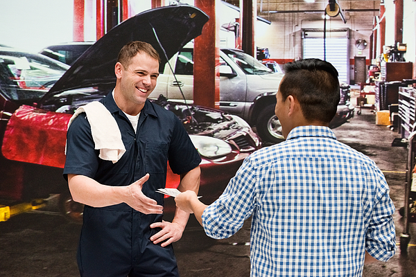 A male customer handing a male mechanic some items in a car repair shop.