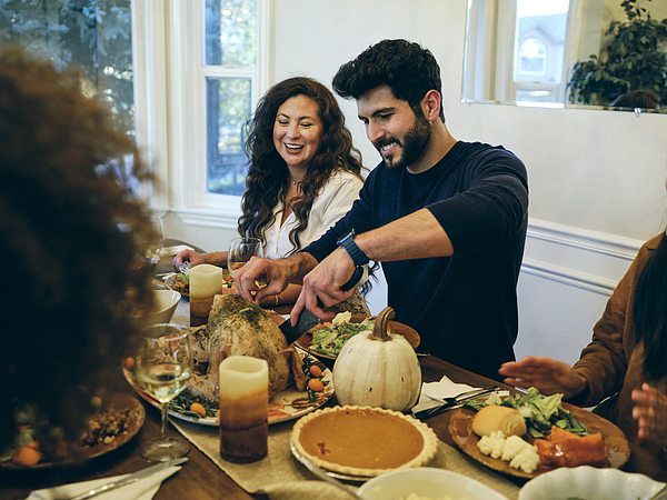 A young man at Thanksgiving lunch cuts into the turkey as a woman looks on, smiling.