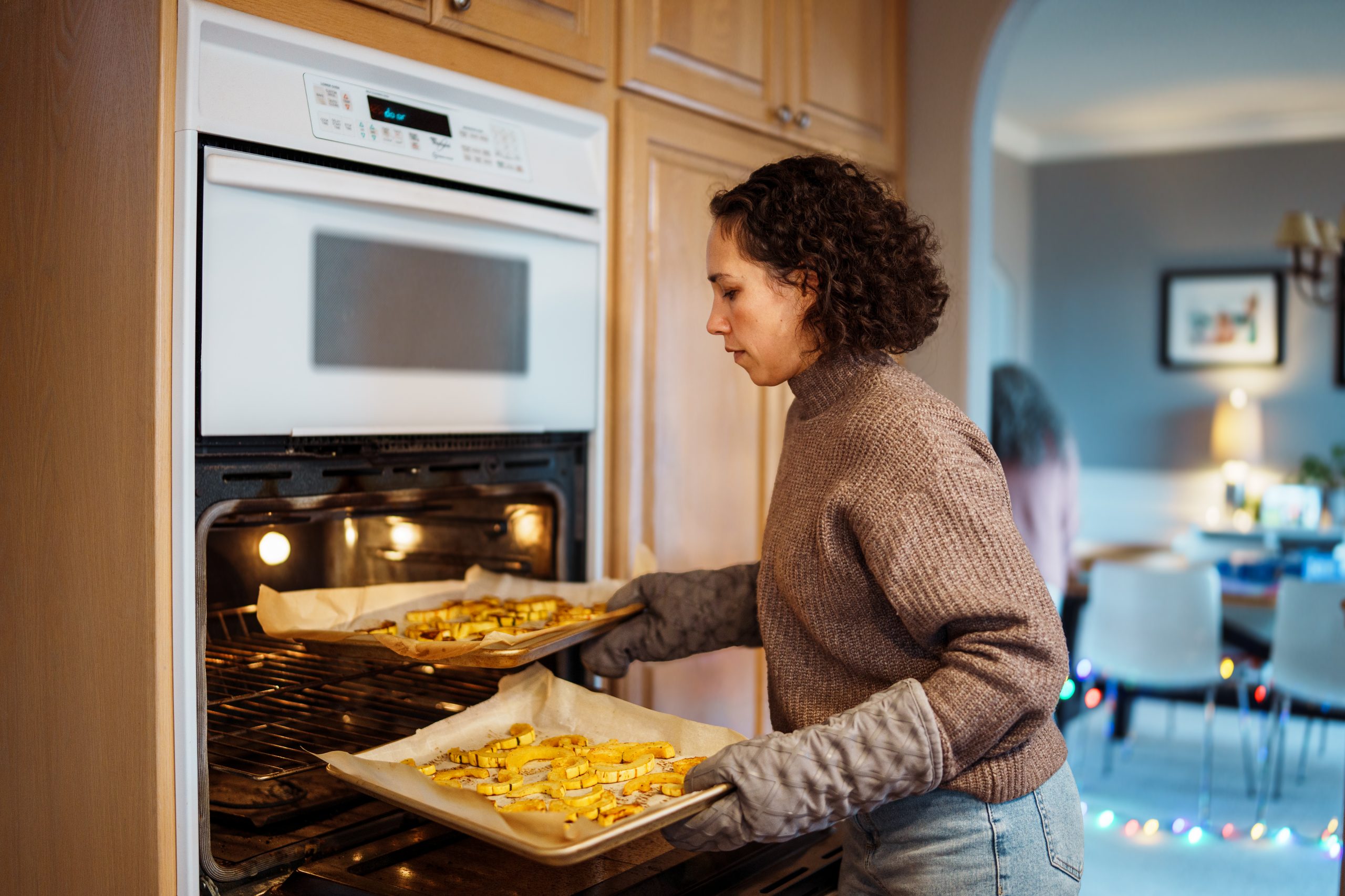 A woman wearing oven mitts removes roasted vegetables from an oven.