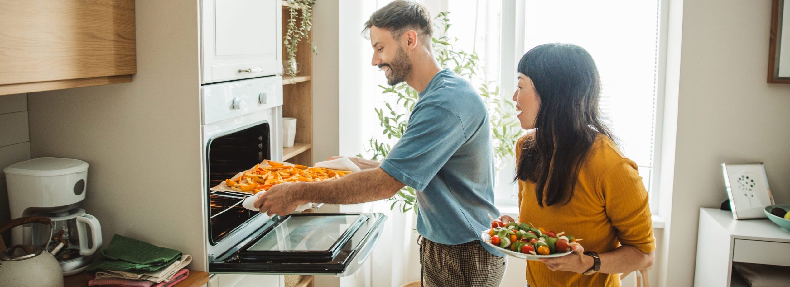 A woman wearing oven mitts removes roasted vegetables from an oven.