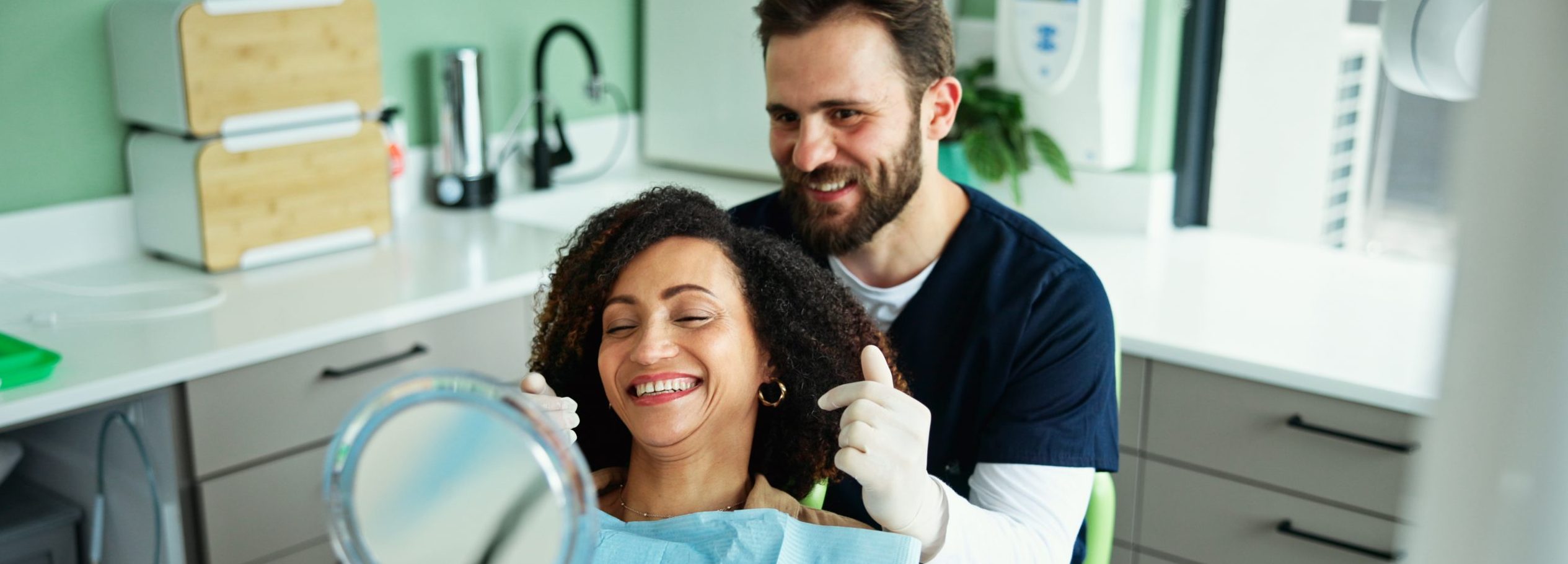 In the dentist’s chair, a woman checks out her smile in a handheld mirror.