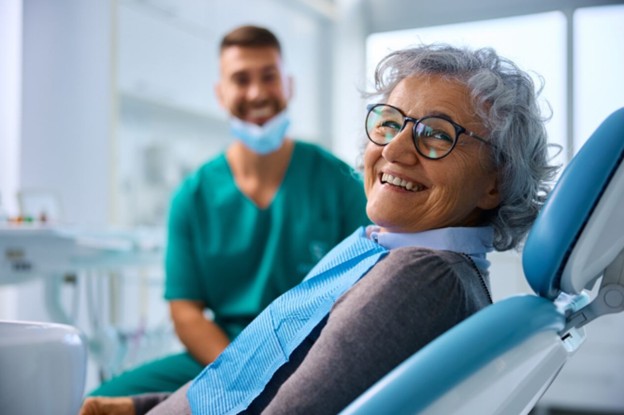 A smiling senior woman sits in a dental chair as a dental hygienist looks on.
