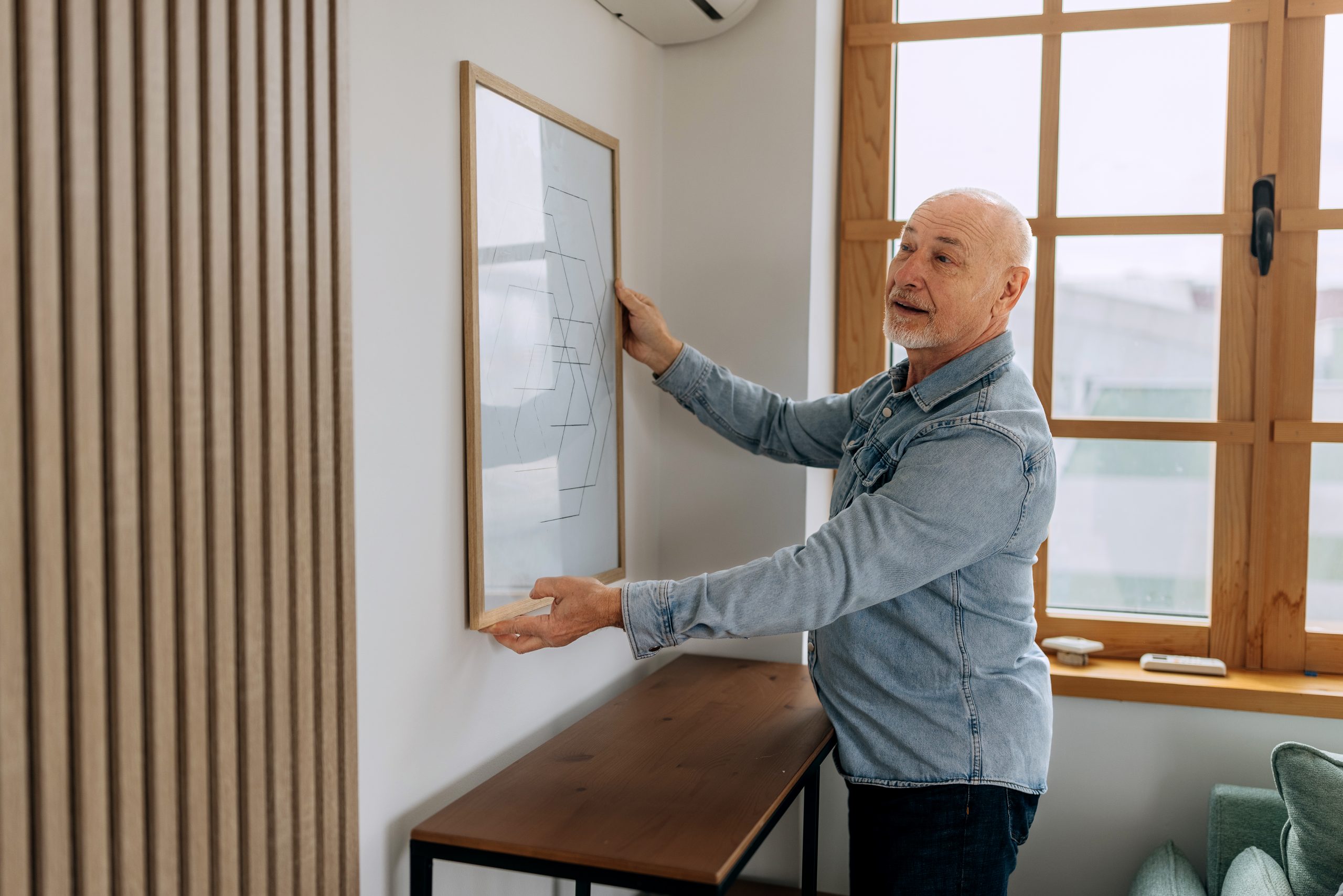 An older man straightens a painting on the wall.