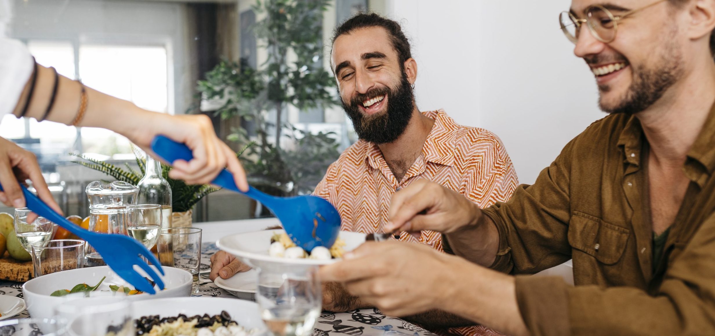 A group of friends gather at the table, laughing and smiling as someone serves them a salad.