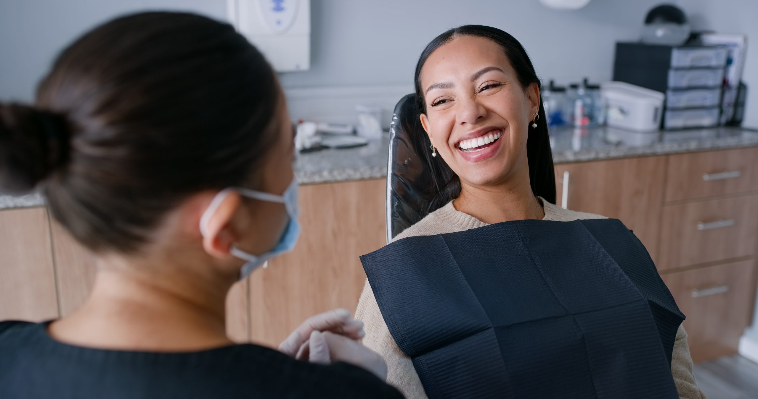 A patient, ready for a dental exam, smiles as a dental hygienist looks on.