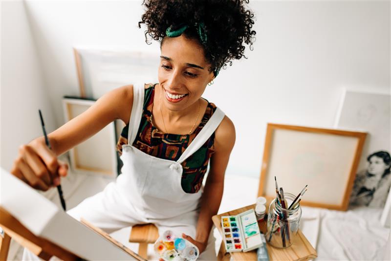 A young woman in overalls paints on a canvas.